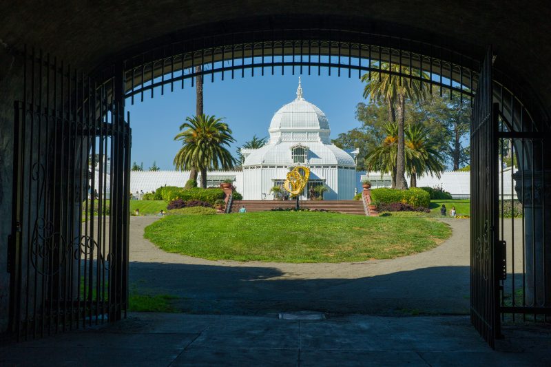 The Conservatory of Flowers framed by entrance gates, palms, and garden greenery in Golden Gate Park.