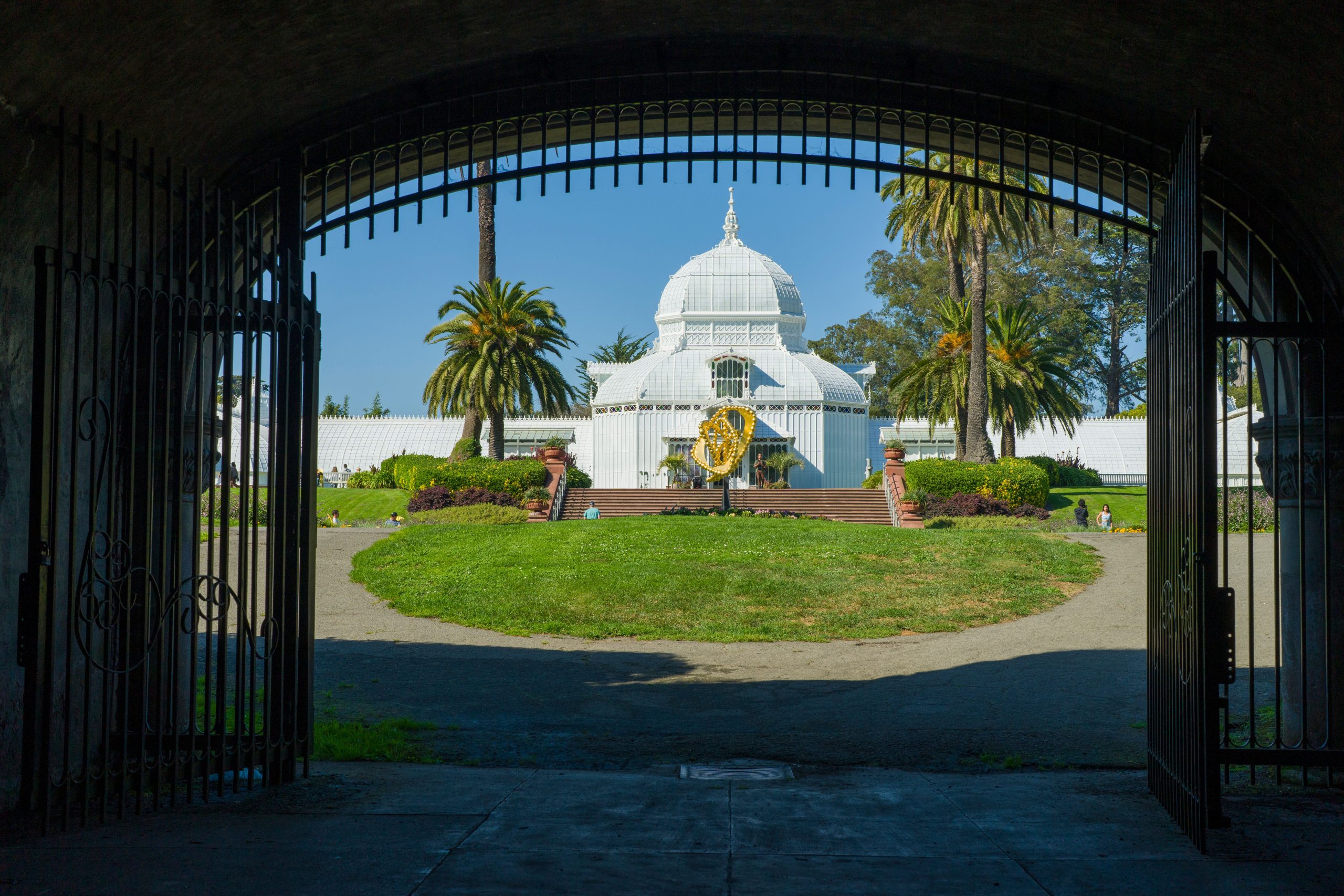 The Conservatory of Flowers framed by entrance gates, palms, and garden greenery in Golden Gate Park.
